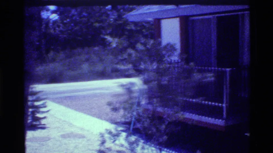 1973 Florida home-movie frame showing a paved path, dense foliage, and a house exterior with balcony railing at frame right.