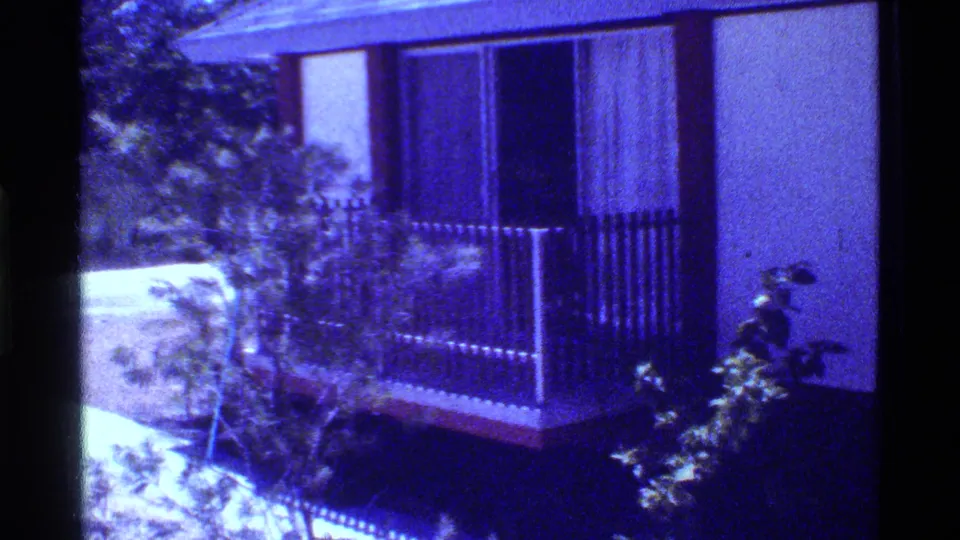 1973 Florida home-movie view of a raised house corner with a dark balcony railing, shrubs, driveway, and trees.