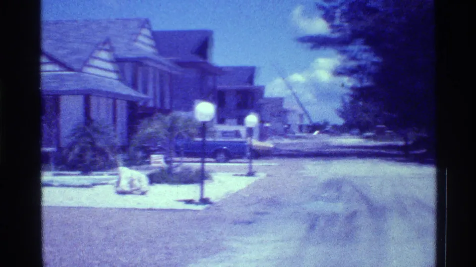 1973 Florida home-movie view down a suburban street with light-sided homes, lawn lamps, trees, and a blue car parked near the curb.