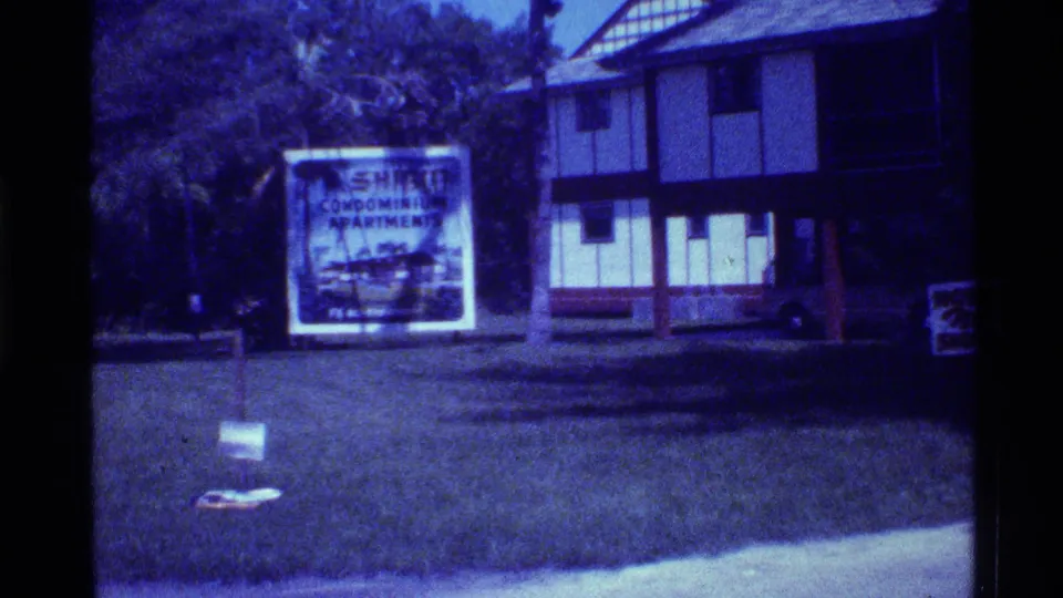 1973 Florida frame showing a Shore condominium apartments billboard on grass near a two-story exterior with trees and a small roadside sign.