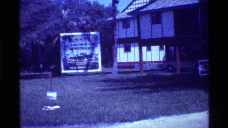 The camera passes a Shore condominium apartments sign in front of grass, trees, and a two-story building with half-timber-like exterior trim.