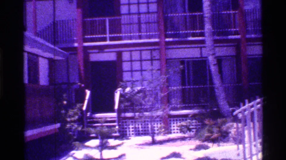 1973 Florida 8mm frame showing an apartment entrance area, balcony railings, gridded windows, palms, and overgrown plants.