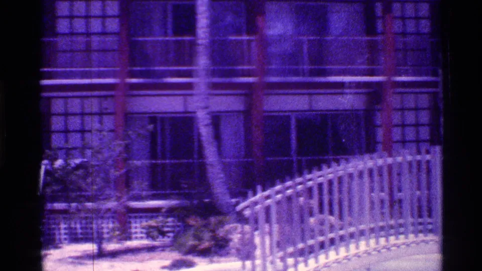 1973 Florida 8mm frame of an apartment exterior with gridded windows, palm trunks, railing, and a curved fence in front.