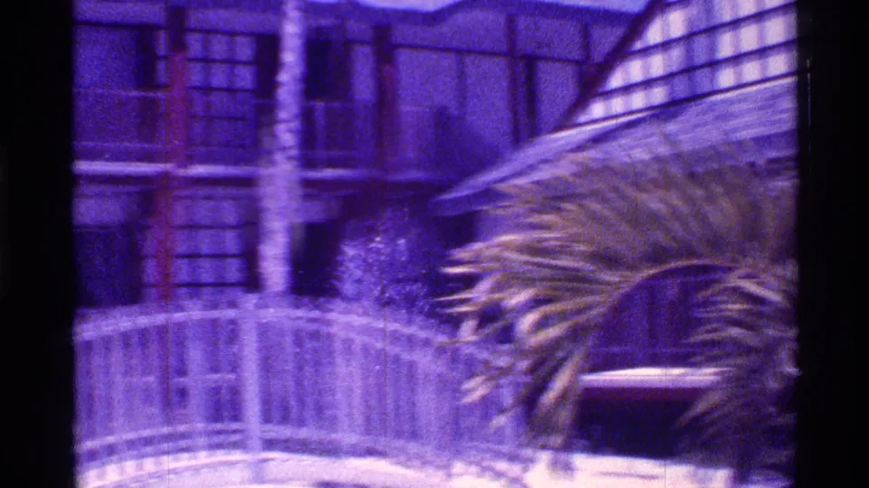 Purple-tinted 8mm frame of a Florida resort exterior with palm fronds, balcony railings, and landscaped grounds.