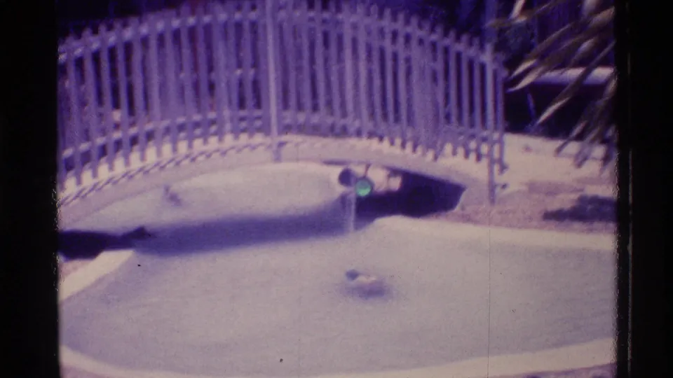 Aged 8mm frame from Florida in 1973 showing a curved white bridge, pale fence, shallow pool, and small fountain spray.