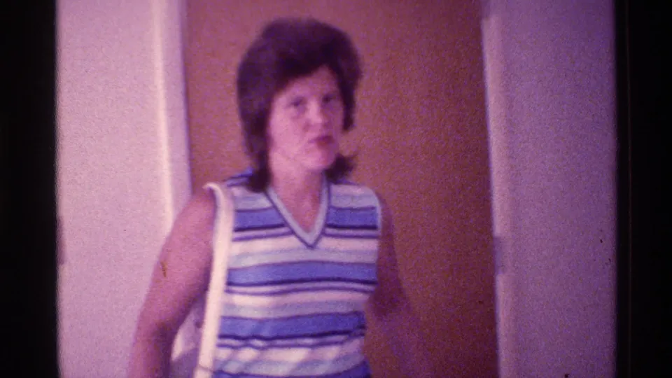 1973 Florida home-movie frame showing a woman with a white shoulder bag moving through a dim doorway in a private interior.