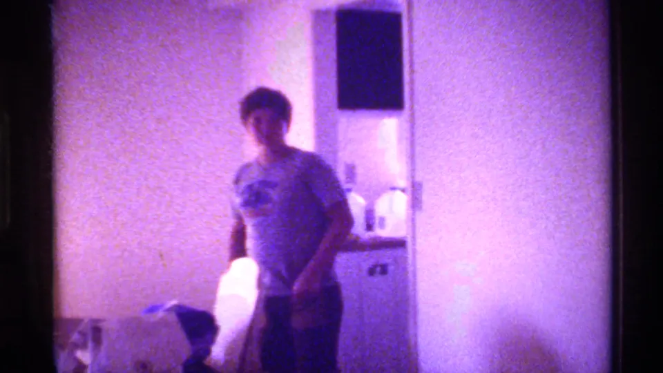 1973 Florida home-movie frame of a young person in a graphic T-shirt standing inside a dim room with a white object nearby.