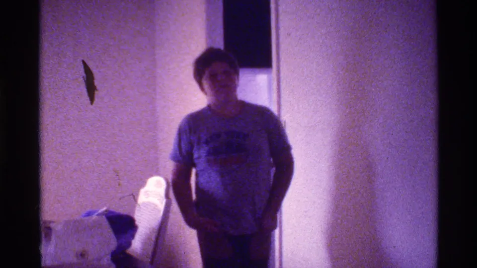 1973 Florida home-movie frame showing a young person in a dim private interior, with a white object on a surface at left.
