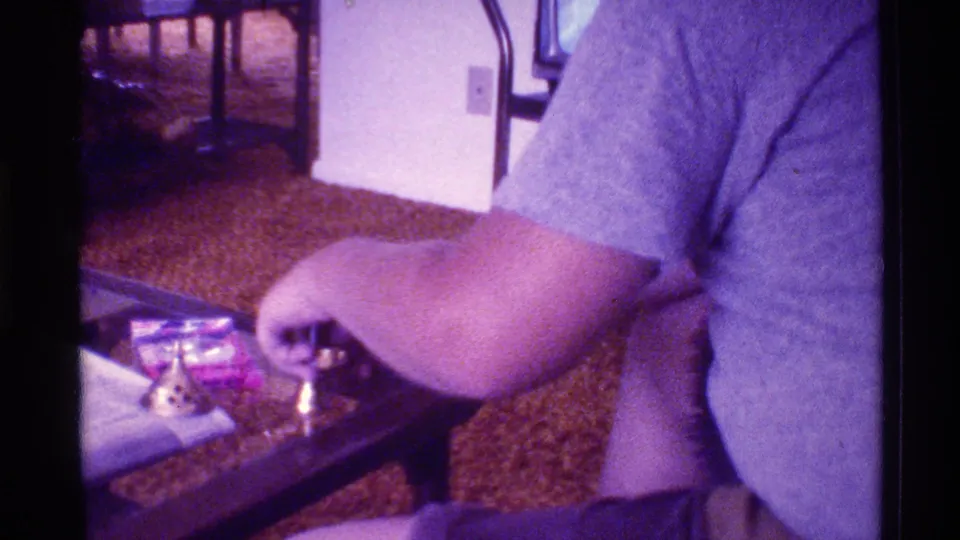 In a Florida 1973 living room, a person in a gray T-shirt handles small items near a brass object on a glass table.