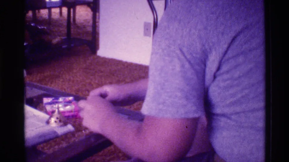 A person in a gray shirt bends over a glass-topped table in a 1973 Florida home interior, with small objects and carpet visible.
