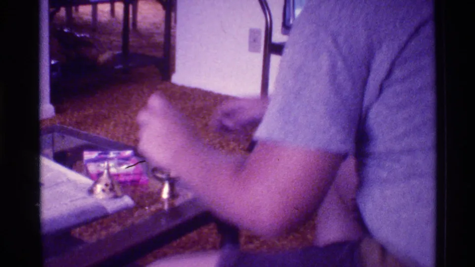 A close 8mm view shows a person working over a glass table in a Florida 1973 living room, with a brass object and small container nearby.