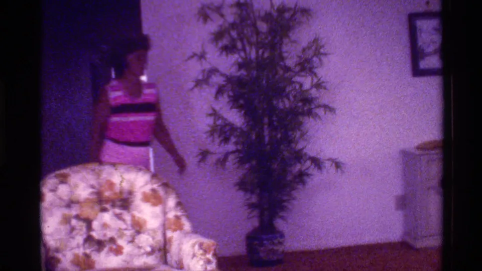 1973 Florida home-movie frame with a woman in a pink and black striped outfit walking past a floral armchair and potted plant.