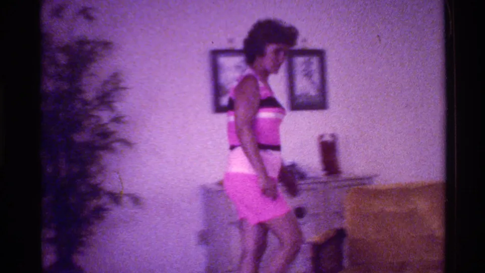 1973 Florida Super 8 frame showing a woman in a pink outfit walking through a living room beside a cabinet, wall art, and chair.