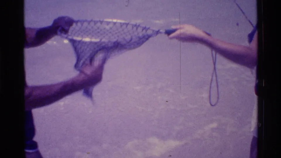 Purple-shifted 8mm beach frame with two partially visible people handling a mesh fishing net in shallow water.