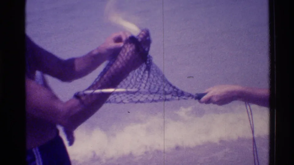 Close beach view of hands and a partial torso working a fish from a mesh net, with foamy surf in the background.
