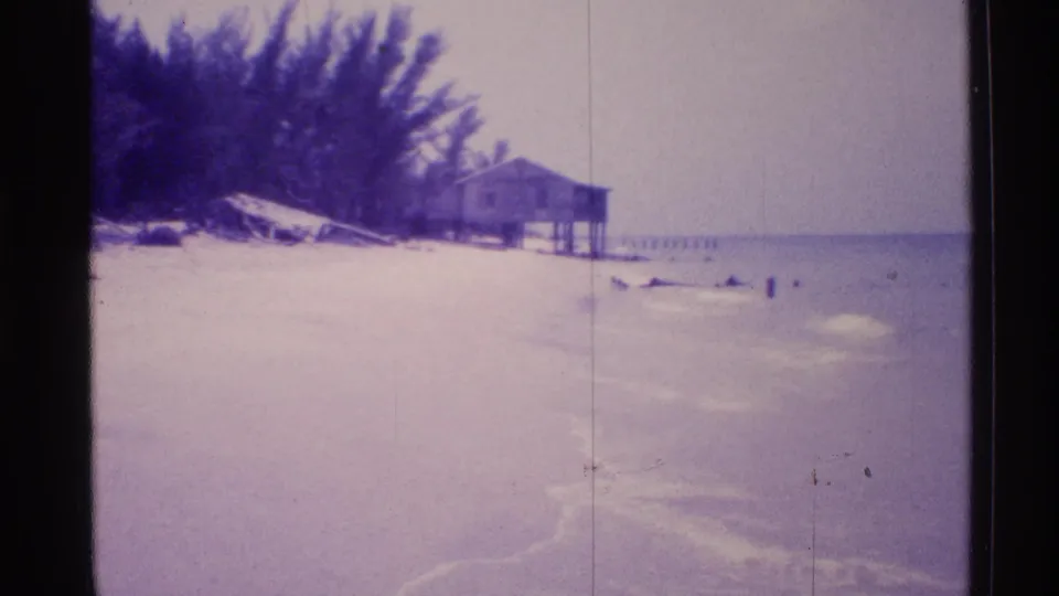 Wide 1973 Florida beach view showing pale sand, surf, trees, and a distant beach house or pier-like structure along the shoreline.