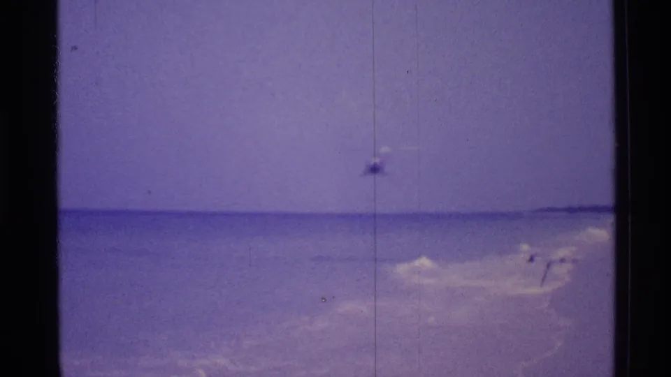1973 8mm Florida beach footage shows a helicopter over open water, softened by purple color shift and vertical film marks.