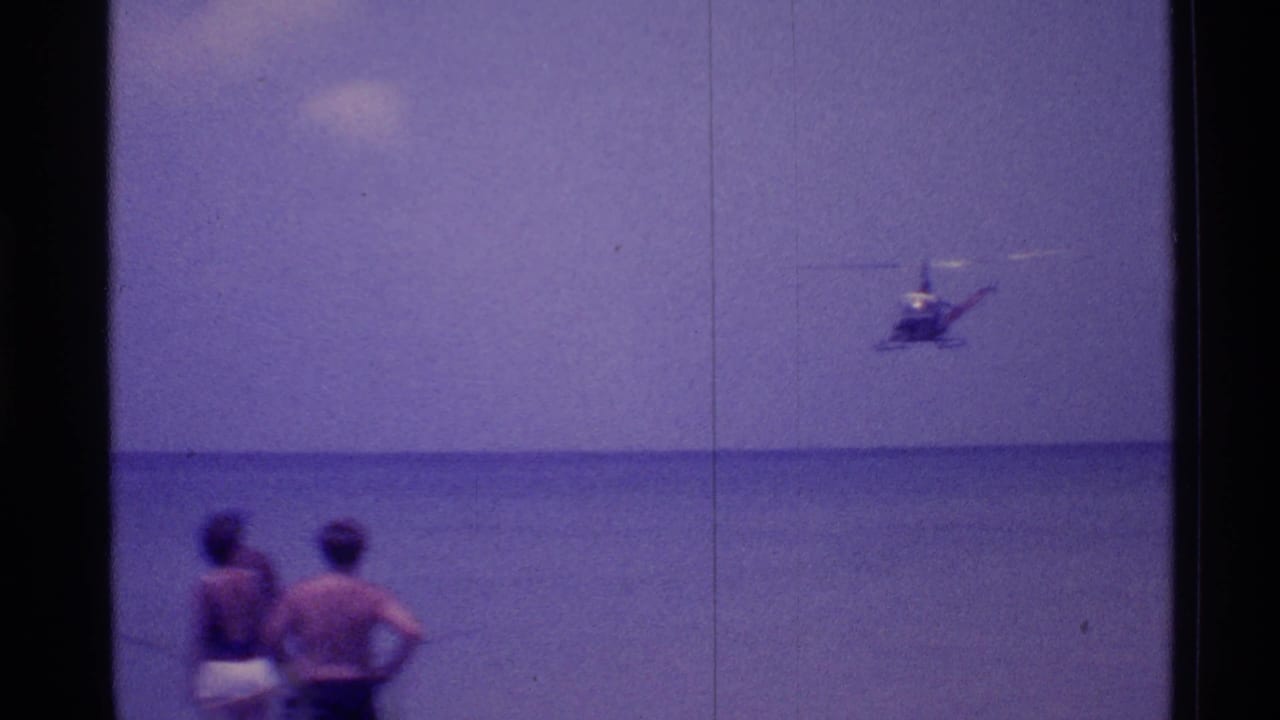 Two beachgoers stand near the water watching a helicopter offshore.
