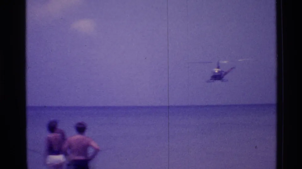 Two people in swimwear stand at the Florida shoreline as a helicopter flies low over the ocean in 1973 home-movie footage.