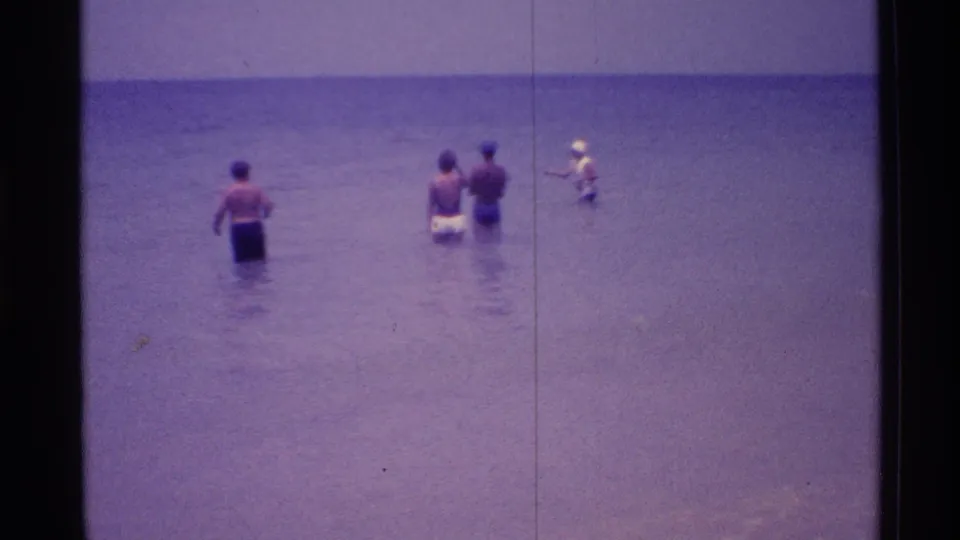 1973 Florida beach frame with adults in shallow surf and a person at right appearing to fish from the water.