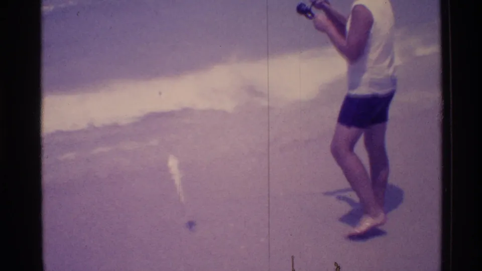 1973 Florida beach frame showing a man on the sand with a handheld camera beside the incoming surf.