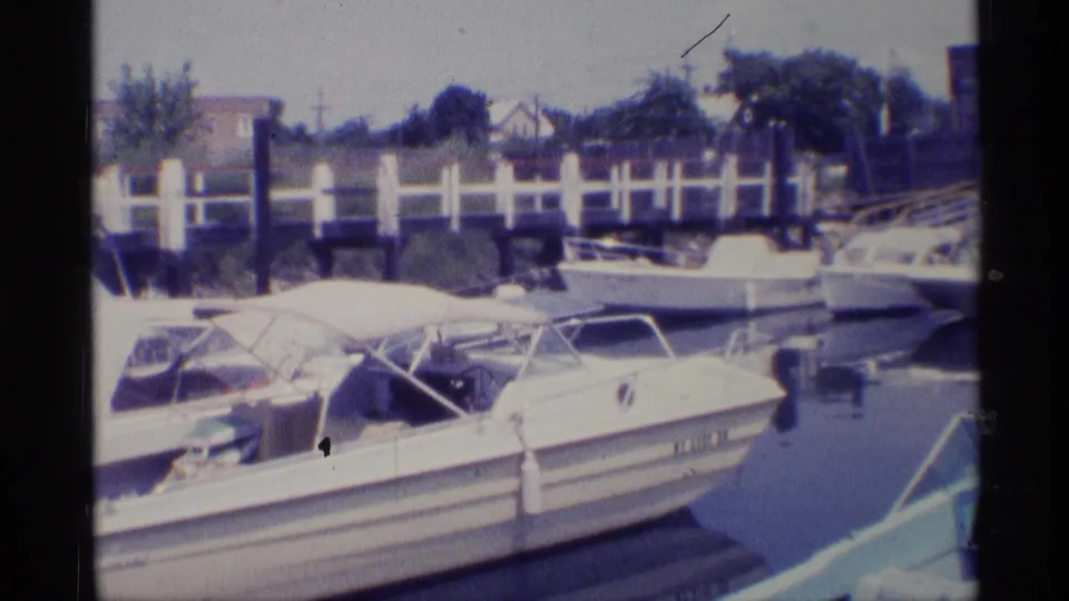 1973 New York home-movie frame of docked motorboats, canopy frames, marina railing, calm water, and distant houses.