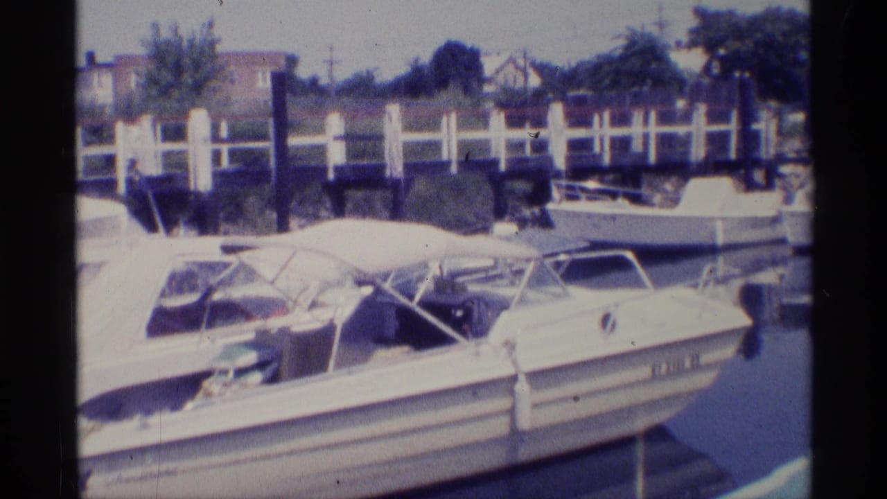 1973 New York 8mm frame showing small docked motorboats, white canopy fabric, dock posts, and residential buildings beyond the marina.