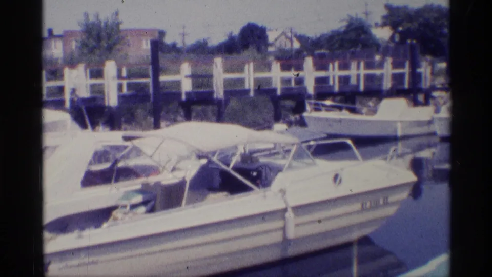 1973 New York 8mm frame showing small docked motorboats, white canopy fabric, dock posts, and residential buildings beyond the marina.