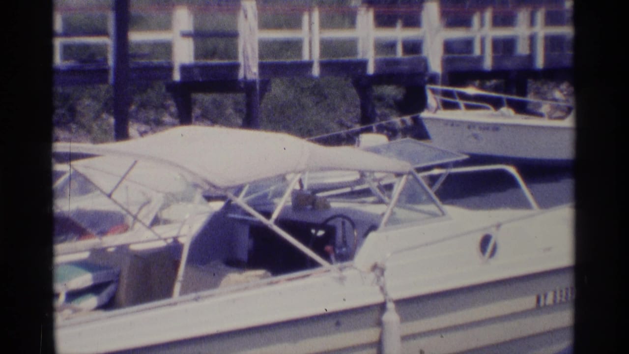 Close 1973 New York 8mm view of a white motorboat with canopy frame, neighboring boats, dock railing, and marina water.