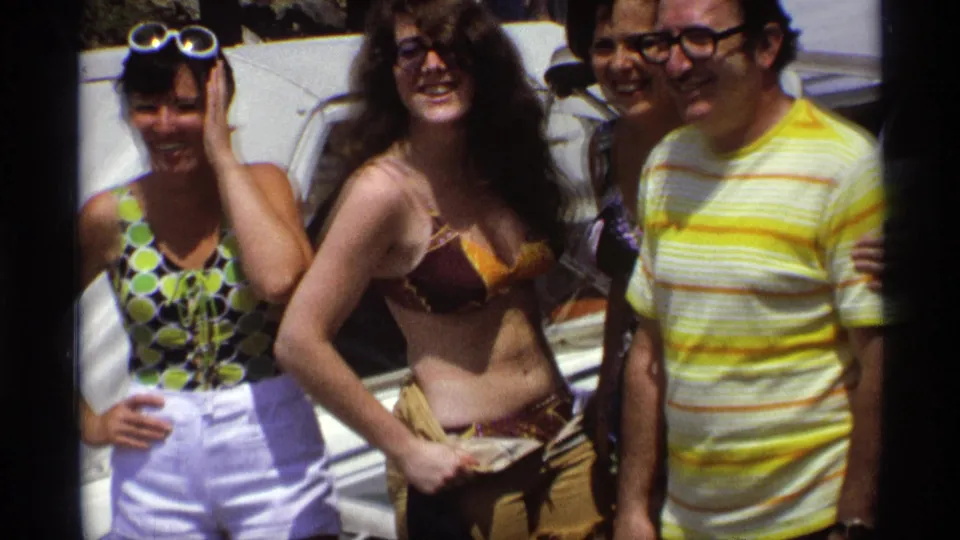 Four adults pose closely on a boat in 1973 New York home-movie footage, with swimsuits, white shorts, and a yellow striped shirt visible.