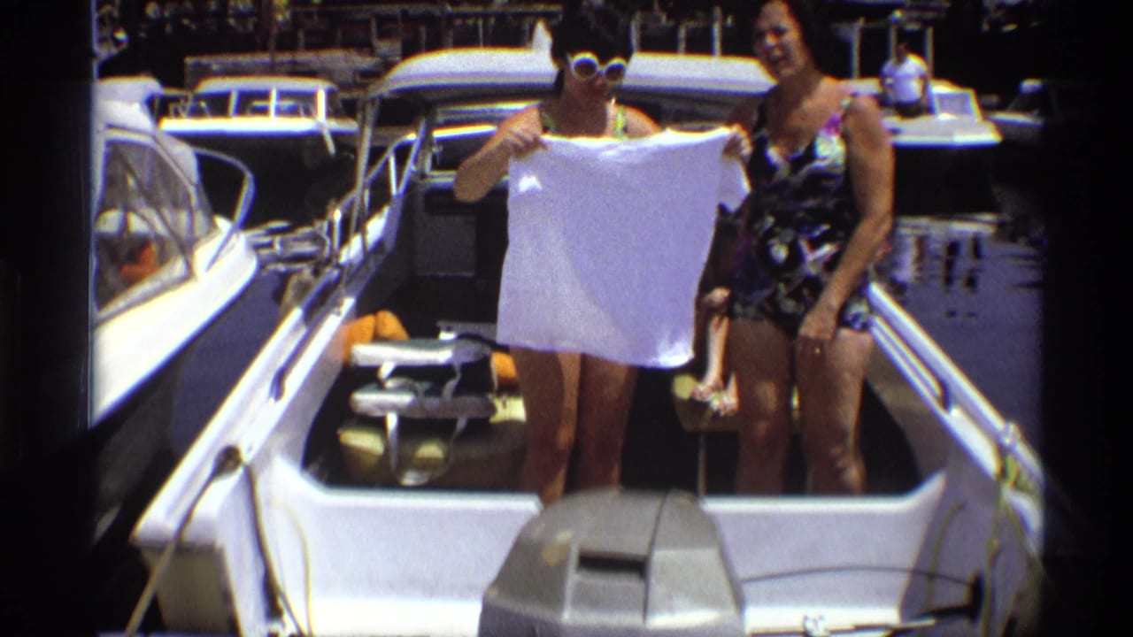 A woman in a patterned swimsuit and white sunglasses holds a white T-shirt on a marina motorboat in 1973 New York footage.