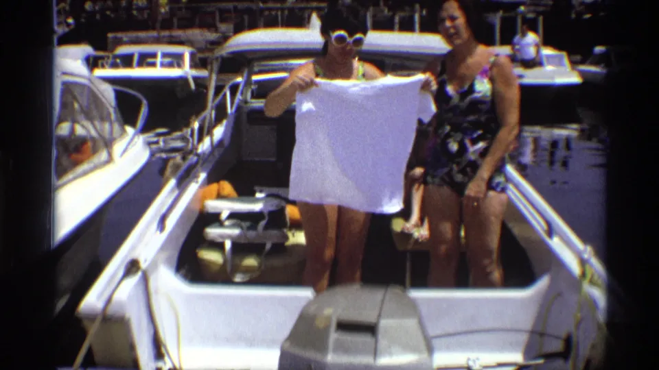 A woman in a patterned swimsuit and white sunglasses holds a white T-shirt on a marina motorboat in 1973 New York footage.