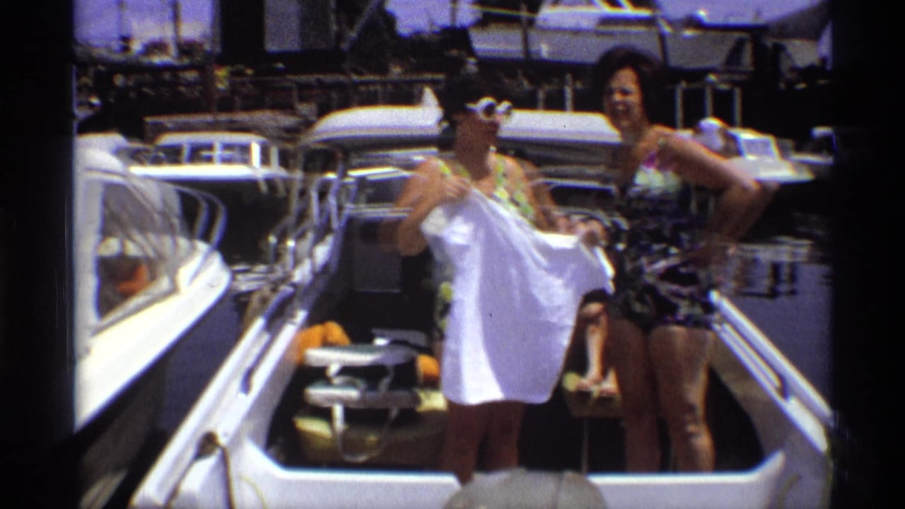 Two women in 1970s swimwear pose on a small boat, with one displaying a white shirt against a marina backdrop.