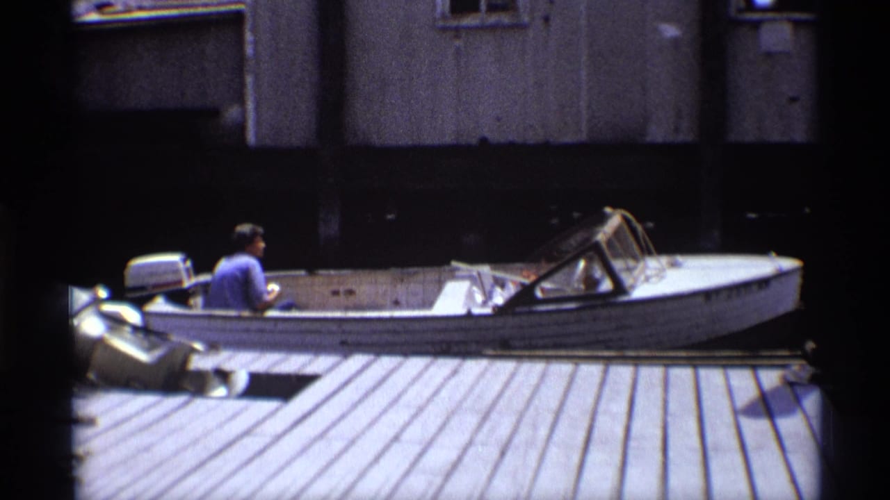 1973 New York 8mm frame showing a man from behind in a blue shirt steering a weathered white motorboat near a planked dock.