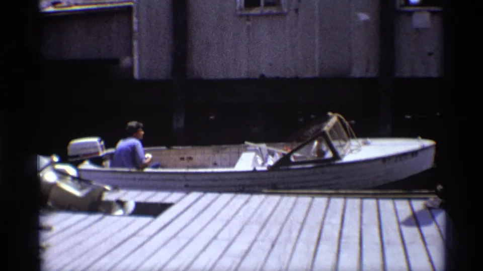 1973 New York 8mm frame showing a man from behind in a blue shirt steering a weathered white motorboat near a planked dock.