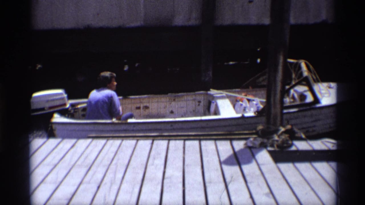 The man in a blue shirt steers the white motorboat past the wooden dock, with ropes and dark waterfront structures behind it.