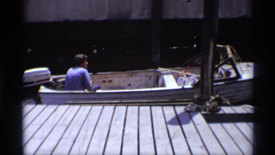The man in a blue shirt steers the white motorboat past the wooden dock, with ropes and dark waterfront structures behind it.