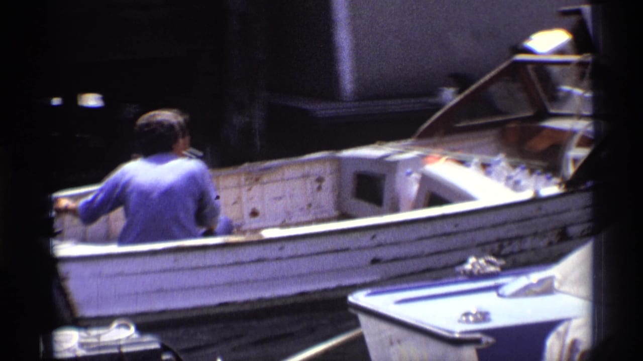 The man continues steering the small white motorboat away from the wooden dock, with another boat edge visible in the foreground.