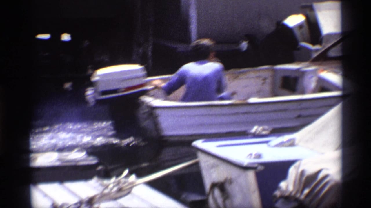1973 New York home-movie frame showing a person seated in a small motorboat at a dock as water ripples nearby.