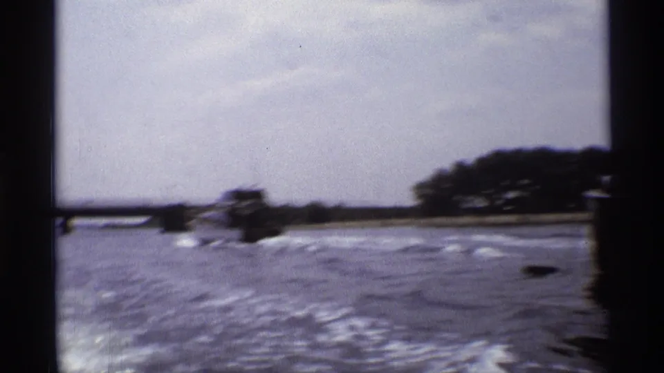 A wide 8mm view shows a small motorboat crossing open river water with shoreline trees in the distance.