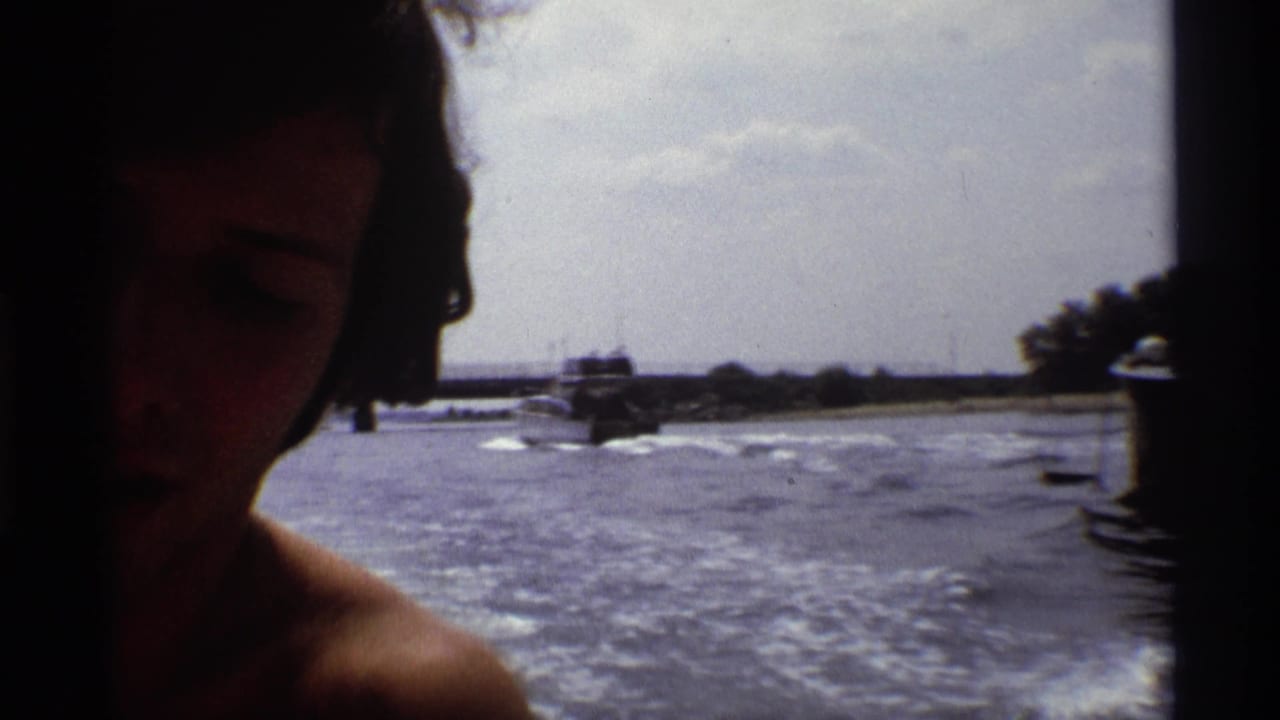 A partially shadowed face appears close to the camera as a small boat continues across the New York river behind them.