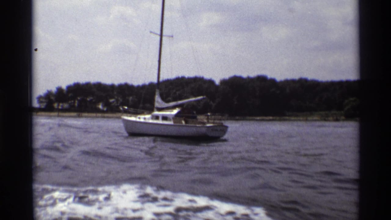 1973 New York 8mm frame showing a white sailboat across lake water, viewed between dark supports from a moving boat.