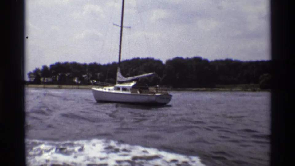 1973 New York 8mm frame showing a white sailboat across lake water, viewed between dark supports from a moving boat.
