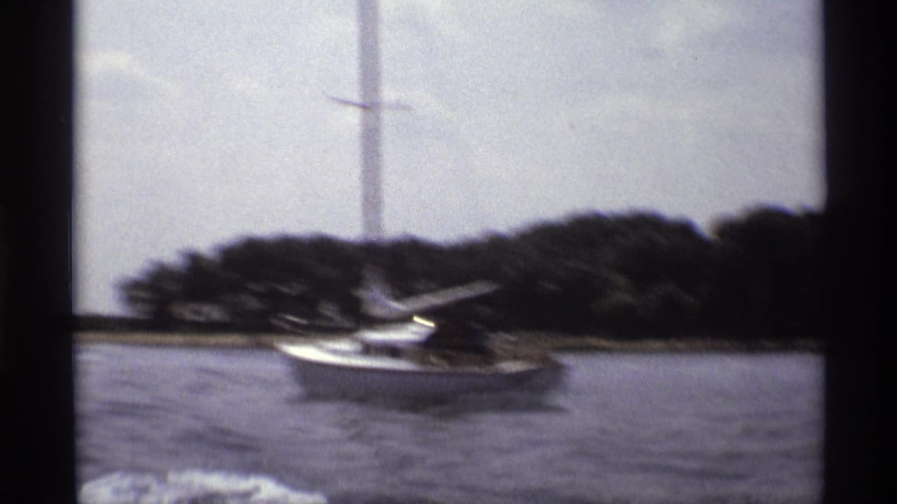 1973 New York 8mm frame with a motion-blurred white sailboat, lake surface, tree-lined shore, and boat wake.