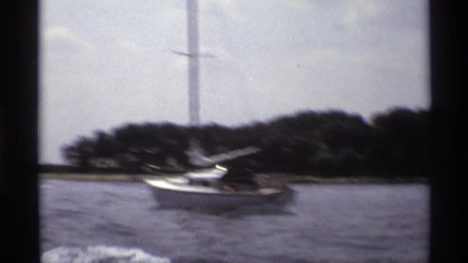 1973 New York 8mm frame with a motion-blurred white sailboat, lake surface, tree-lined shore, and boat wake.