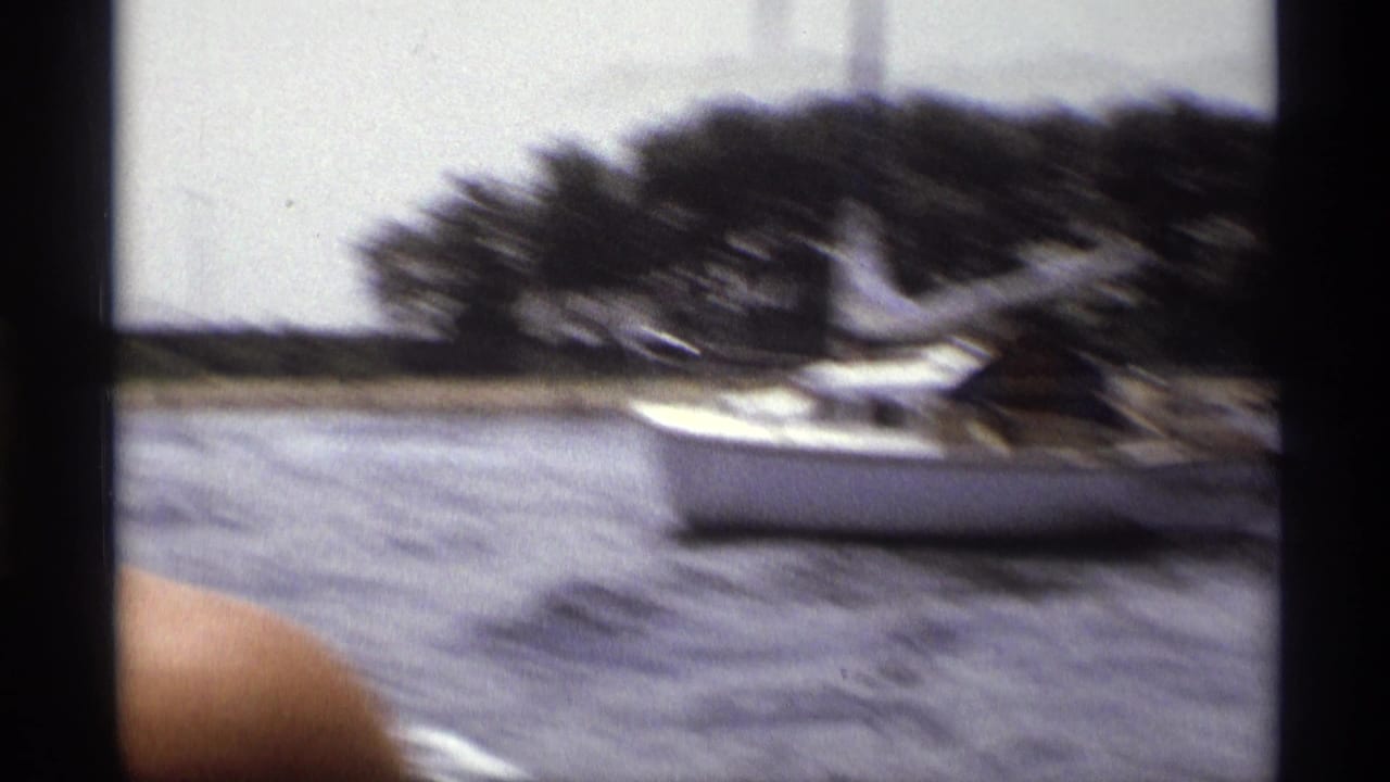 1973 New York 8mm frame of a sailboat near a tree-lined shore, with lake water and motion blur from a moving boat viewpoint.
