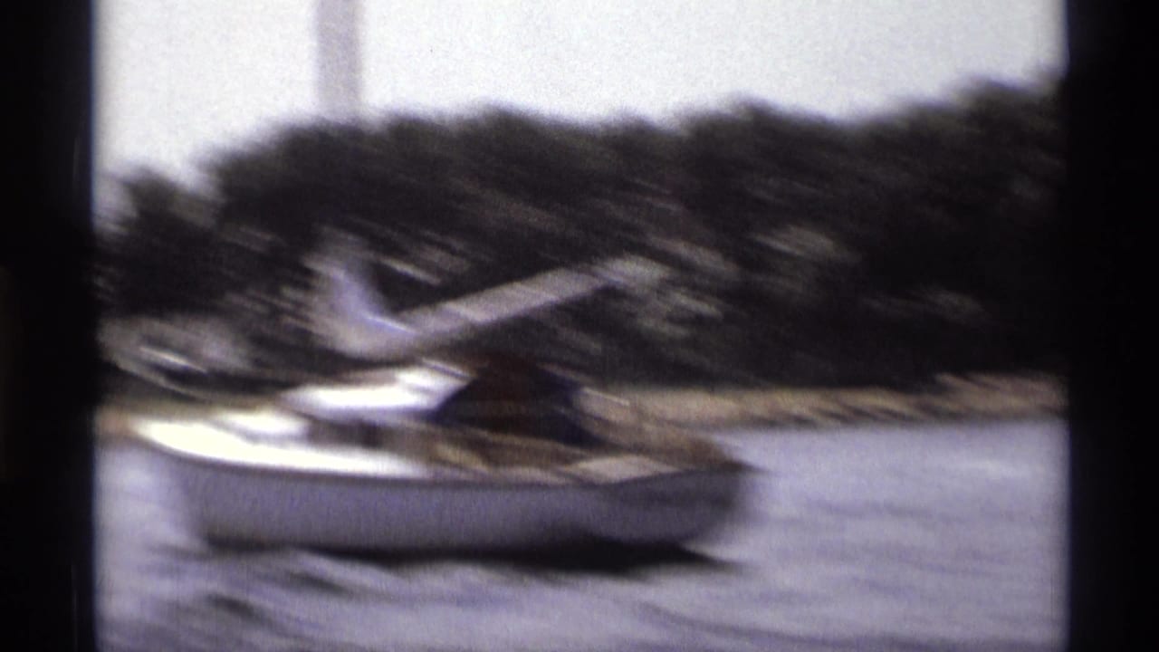 Blurred sailboat crossing river water near a wooded shoreline.
