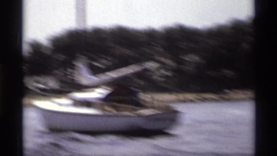 Vintage 8mm frame of a sailboat moving across a New York river, with trees along the shore and motion blur from the camera boat.
