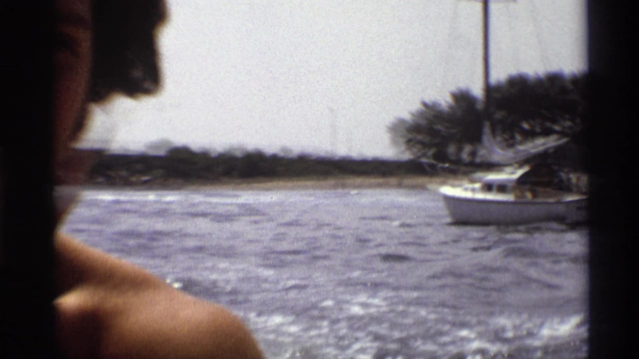 A 1973 New York boating view shows an adult passenger partly in frame, open water, and a white sailboat near the wooded shoreline.