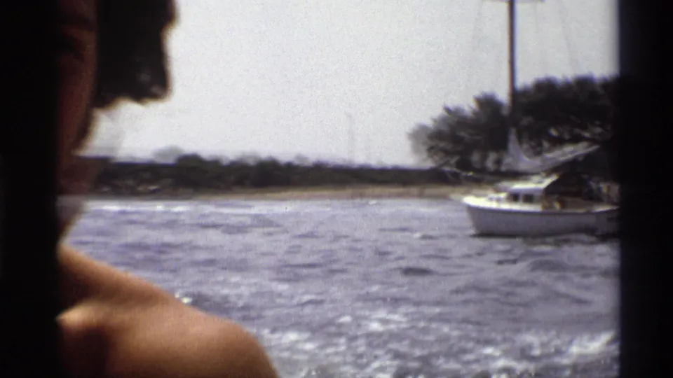 A 1973 New York boating view shows an adult passenger partly in frame, open water, and a white sailboat near the wooded shoreline.